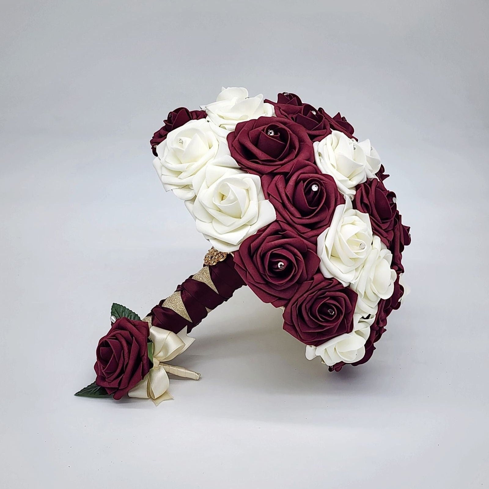 Burgundy and ivory wedding bouquet made with Real Touch Roses. Rhinestones are in the center of Roses. Handles is ivory and burgundy French twist and finished withgold brooch and gold bling wrap