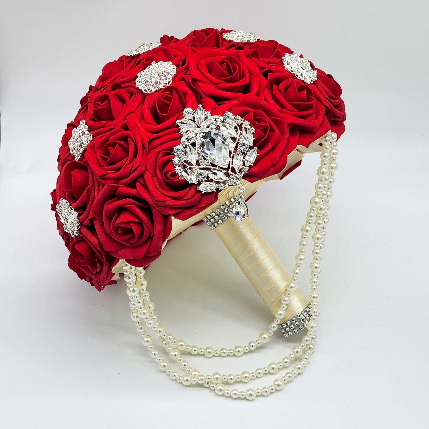 Red bridal bouquet with cascading pearls. Brooches are scattered throughout bouquet. Handle is made using ivory satin ribbon. Finished with silver bling wrap